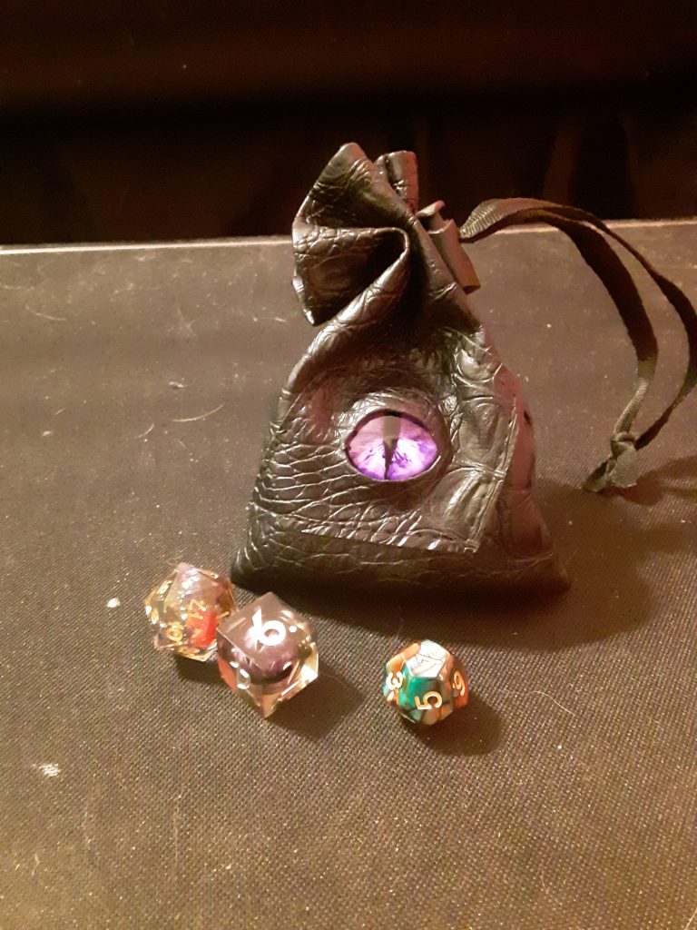 A leather dice bag with an eye in the center next to 3 die.